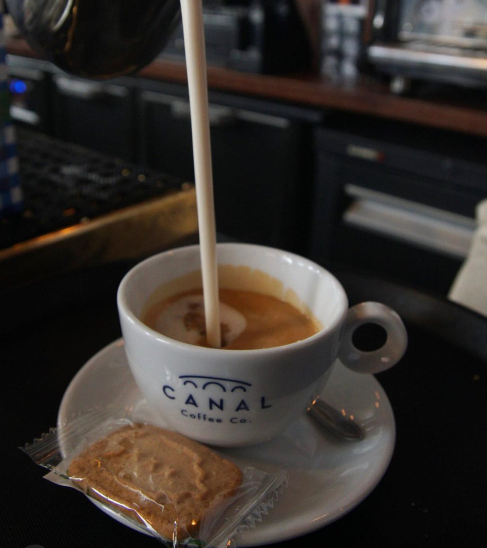 Melk wordt ingeschonken in een Canal Coffee Co. kopje espresso met een koekje op het schoteltje.