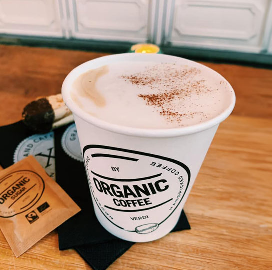 Biologische cappuccino geserveerd in een duurzame beker, afgetopt met een vleugje kaneel en een koekje voor erbij.







